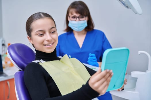 Happy young teenage female with mirror sitting in dentist chair smiling looking at healthy teeth, with dentist doctor in office. Dentistry, hygiene, treatment, dental health care concept