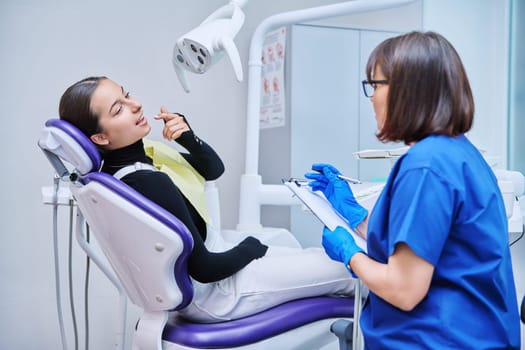 Dentist and teenage girl patient in dental clinic, doctor nurse writing on clipboard consulting, teen female talking about toothache disturbing dental problem. Dentistry, treatment, dental health care