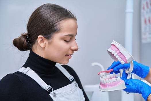 Teenage female sitting in dental chair at dentist checkup, doctor with dental jaw model and toothbrush telling and showing teenager dental care. Adolescence hygiene treatment dental teeth health care