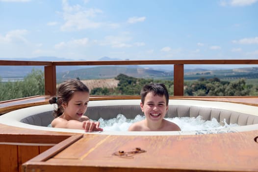 Happy Caucasian kids, a teenage boy and his little sister smiling looking at camera while playing in the swimming pool with bubbles, enjoying happy family holidays in Spain. People. Vacations