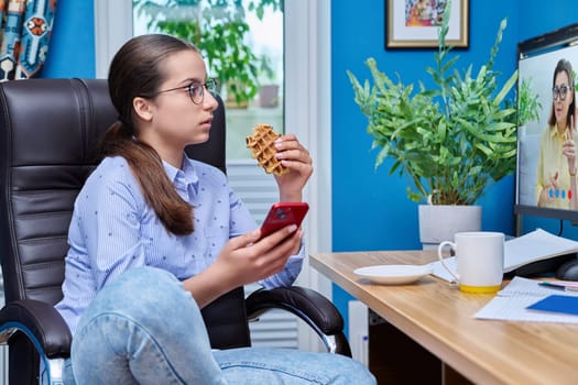 Teenage girl watching a training course sitting at a table at home snacking on waffles. Lifestyle, adolescence, leisure, learning concept