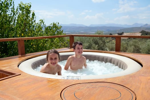 Cheerful teenage boy and school age little child girl, a brother and sister smiling at camera, having fun in the swimming pool, enjoying happy Spanish family holidays. People. Travel Destination.