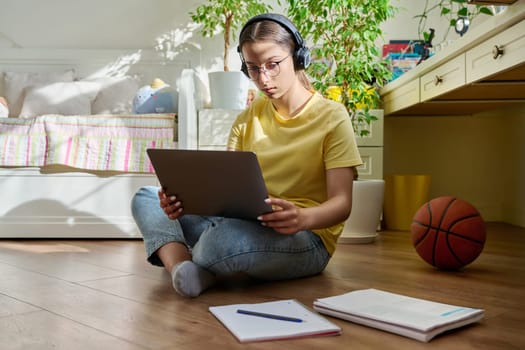 Teenage girl with glasses studying at home using laptop. Female with headphones and textbooks sitting on floor in room. Education, adolescence, high school concept