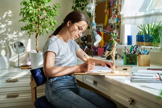 Teenage girl drawing sketches with a pencil, sitting at the table at home. Creative youth, education, learning, hobbies and leisure, adolescence concept