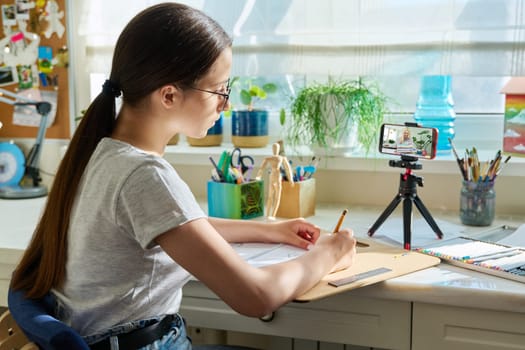 Teenage girl drawing sketch, looking at screen of smartphone, sitting at home at desk, learning an online course in drawing. Education creativity leisure hobby adolescence concept