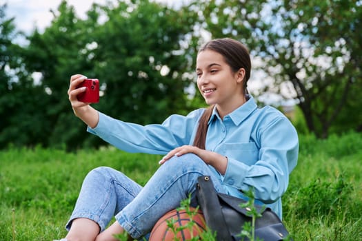 Teenage female student resting sitting on grass using smartphone for video call chat conference. Attractive teen girl on lawn with backpack. High school applications technology lifestyle adolescence