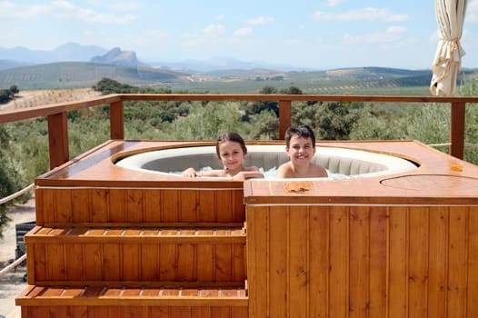 Happy kids, a teenage boy and his little sister smiling looking at camera while playing in the swimming pool with bubbles, enjoying happy family holidays, against the background of huge mountains