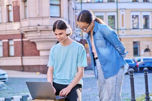 Teenage male and female using laptop for study, leisure, outdoor on city street. Guy and girl teenagers looking together at aptop screen. Lifestyle, technology, youth, education, city life concept