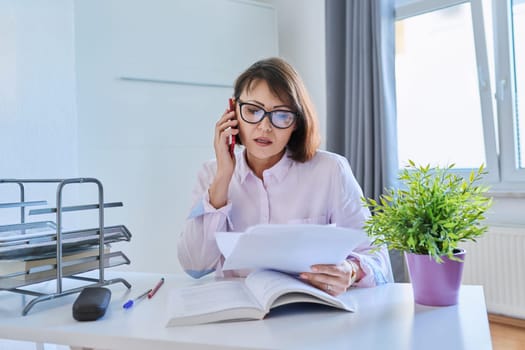 Middle aged business woman working at home sitting at the table with smartphone documents books. Freelance, remote work, home office concept