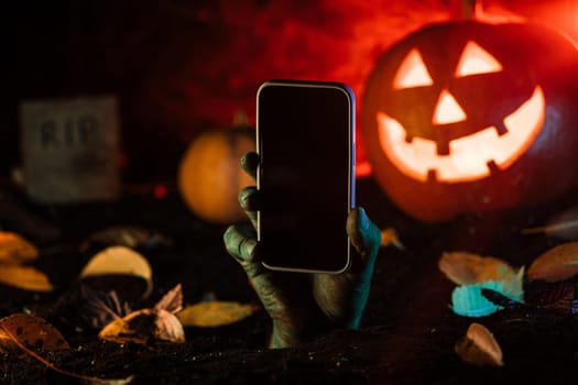 Zombie hand with smartphone. Undead, halloween pumpkin, mobile operator advertising. Terrifying scene from underground. Copy space. High quality