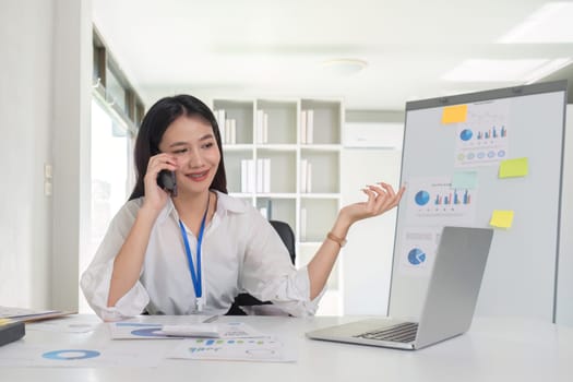 Businesswoman talking on the phone talking with customers and using laptop to do financial management and calculations on white table in office.