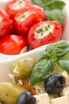 Salad of black, green olives with pieces of cheese. Garnished with basil leaves. The white bowl. Spicy round red peppers stuffed with cheese in the background.