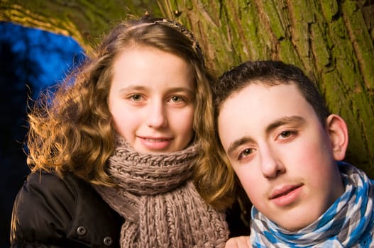 Head and shoulders portraits of two young teenage siblings (brother and sister) standing by a tree at dusk and looking at the camera