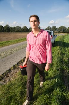 young man walks along a remote country road with a gasoline canister towards the camera  Background of parked car