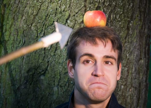 Head and shoulders shot of a young man at the tree with apple on his head and arrow at him from the camera