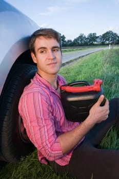 young man sits on the side of the road in the grass at the front wheel of a silver-gray car and handles a black gasoline canister