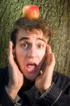 Head and shoulders shot of a young man at the tree with apple on his head and hands on his cheeks looking at the camera anxiously