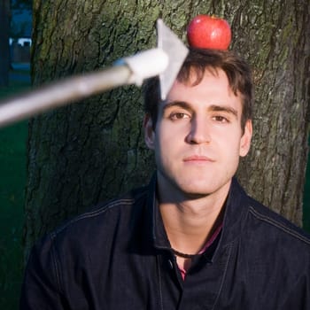 Head and shoulders shot of a young man at the tree with apple on his head and arrow at him from the camera