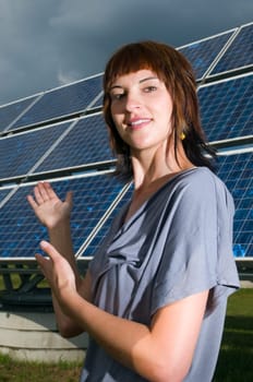 Upper body shot of a young, brunette woman in a casual leisure dress on a green meadow in summer, presenting to a photovoltaic system behind her.