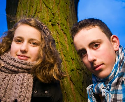 Head and shoulders portraits of two young teenage siblings (brother and sister) standing by a tree at dusk and looking at the camera