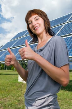 Upper body shot of a young, brunette woman in a casual leisure dress on a green meadow in summer, presenting to a photovoltaic system behind her.