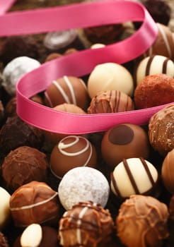 A decorative pink ribbon lies twirled over handmade spherical chocolates with different patterns in a gift and celebration concept