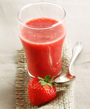Glass of freshly blended strawberry smoothie on burlap with a luscious ripe red strawberry and silver spoon
