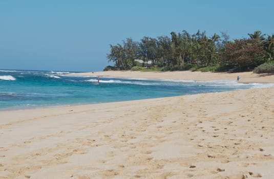 Idyllic tropical sandy beach and bay with a lone surfer in the distance amongst the breaking waves.