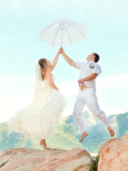 Bride and groom jumping on a rock with umbrella