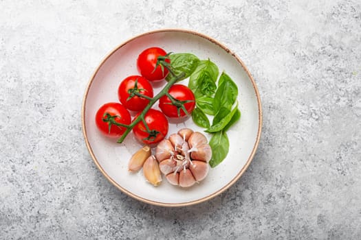 Italian food cooking concept with cherry tomatoes, fresh green basil and garlic cloves top view on plate on white stone background, ingredients for preparing Mediterranean meal.