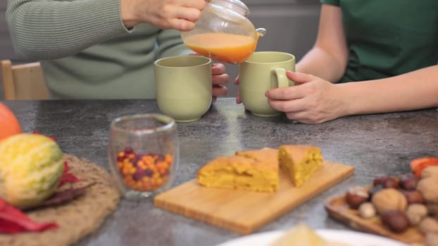 unrecognizable female couple have breakfast together drink orange berry fruit tea with slices of pumpkin pie sitting gray kitchen table celebrating autumn holidays. two women chatting in kitchen