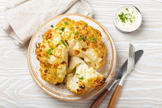 Whole roasted head of cauliflower with olive oil on plate on white wooden rustic table top view with cutlery. Vegetarian dish..