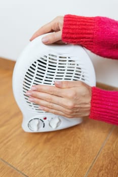 A woman in a plaid and a warm sweater is warming herself near an electric fan heater at home, close-up