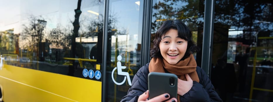 Enthusiastic asian woman, standing on bus stop with smartphone, looking at phone screen with amazed, triumphing face, winning, hear great news on video chat.