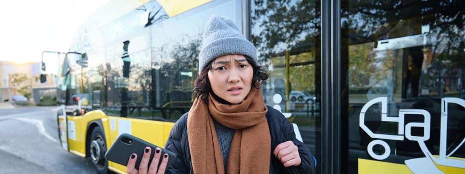 Portrait of confused asian girl, standing on bus stop, holding mobile phone, looking shocked and upset, disappointed by public transport delay.