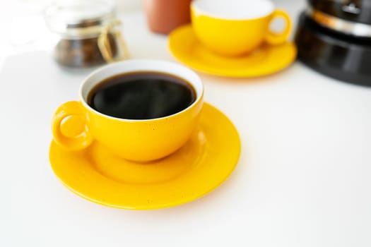 Hot aromatic black coffee in a bright yellow cup and saucer stands on a white table against the background of a coffee maker and a jar of coffee beans. Place for an inscription