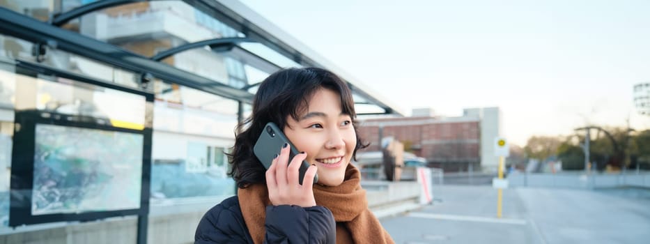 Smilling Korean girl talking on mobile phone, standing on bus stop, using smartphone, posing on road in winter, wrapped in scarf, wearing black jacket.