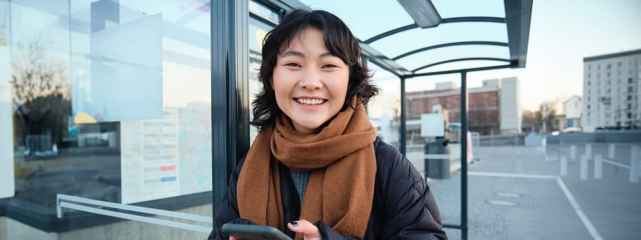 Beautiful young woman, standing on bus stop, waiting for her public transport, using mobile phone, using smartphone application.