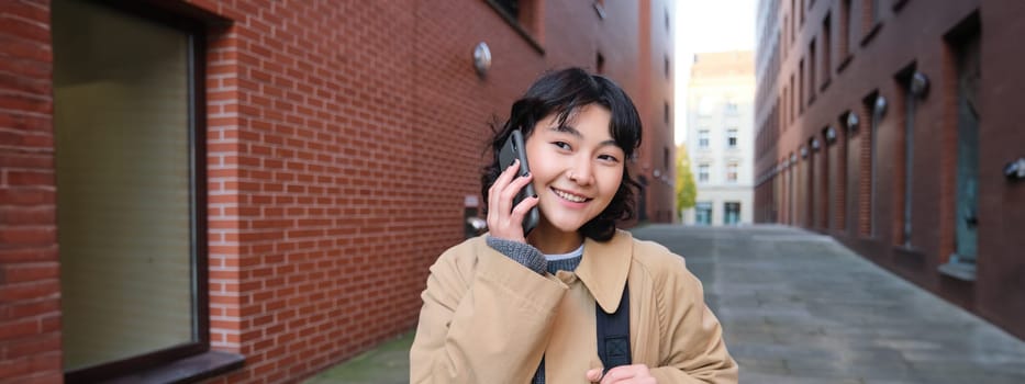 Cellular technology concept. Young modern girl talks on mobile phone, walks on street and smiles, has conversation over telephone. Copy space