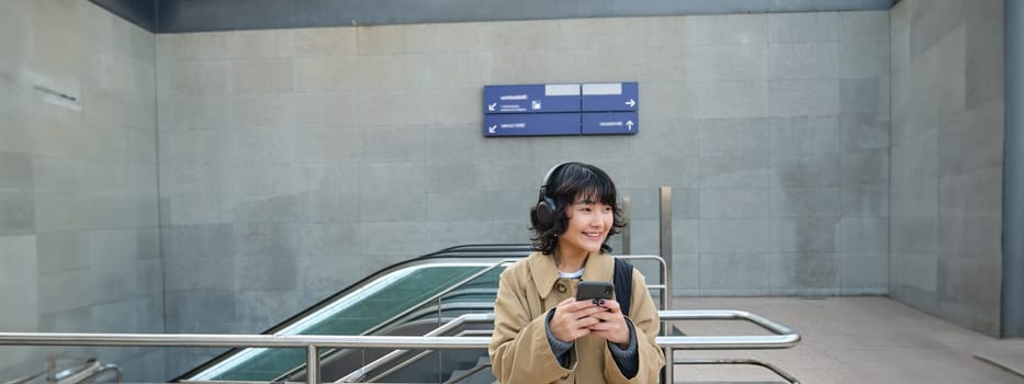 Portrait of stylish asian hipster girl, listens music in headphones, looks at her phone, tourist looking at map for a way to sightseeing, drinking coffee to go.