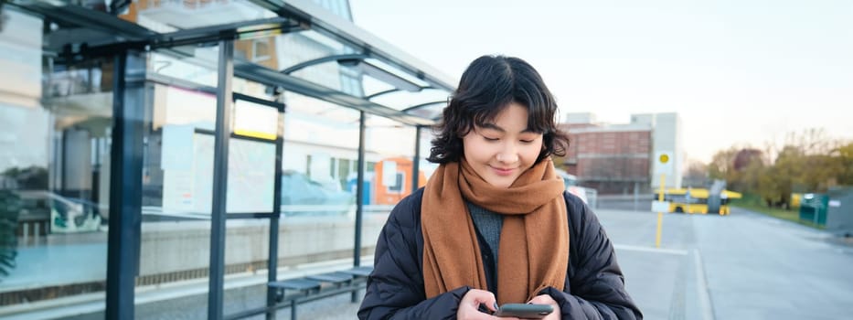 Happy smiling korean girl, using mobile phone, standing on bus stop with smartphone, looking at departure schedule on application, posing in winter clothes.