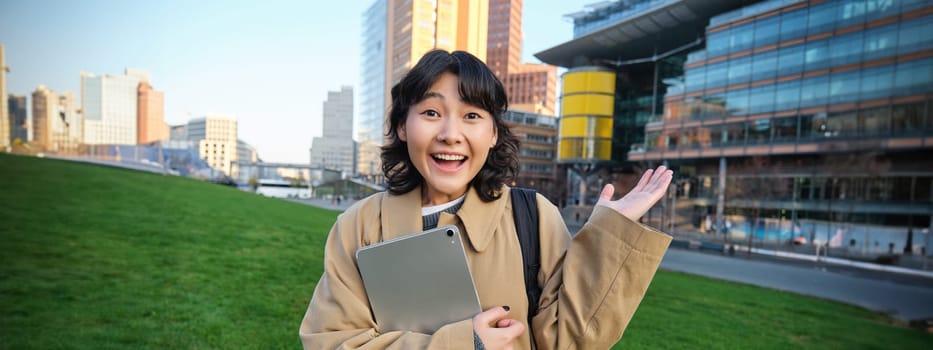Enthusiastic brunette asian girl, student with digital tablet in her hands, looks impressed and surprised, stares at camera with amazement, stands on street in awe.