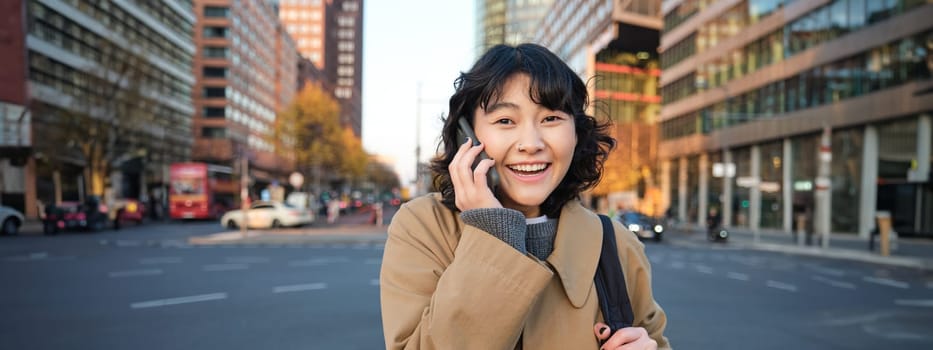 Portrait of happy korean girl, talks on mobile phone, looks surprised and happy, receives positive great news over telephone conversation, stands on street of city.