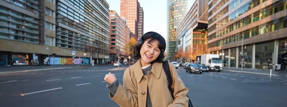 Portrait of young asian girl, student walks in city, listens music in headphones and uses mobile phone on streets, celebrates, triumphs and makes joyful fist pump.