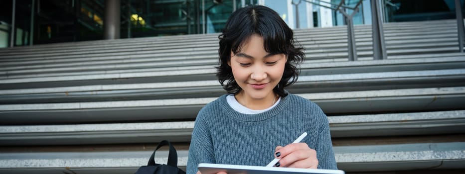 Portrait of asian girl student, hipster sitting on stairs with digital tablet and cup of coffee, draws digital art, makes design project.