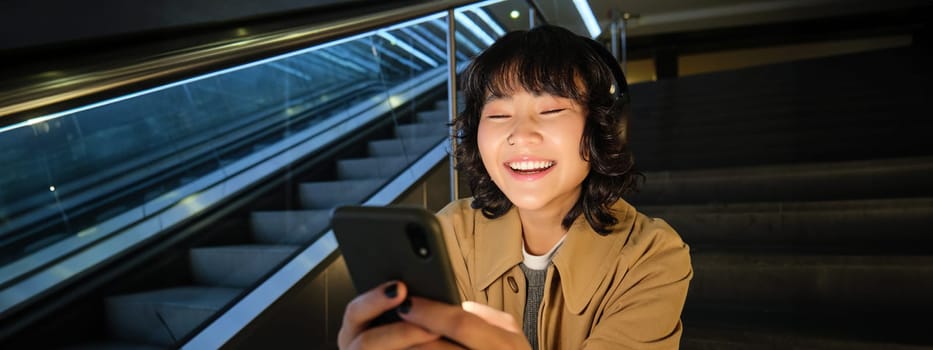 Close up portrait of smiling asian girl student, listens music in headphones and looks at mobile phone, uses smartphone, sits on staircase in public place.