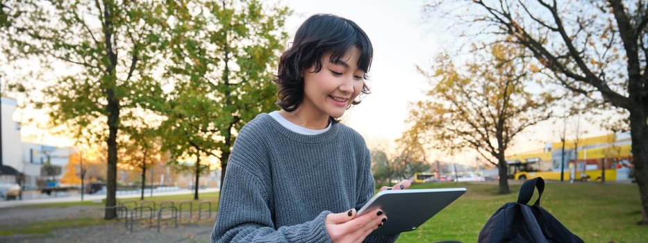 Portrait of young smiling korean girl, graphic designer, artist drawing on digital tablet with a pen tool, sitting in park on fresh air and scatching, taking notes.