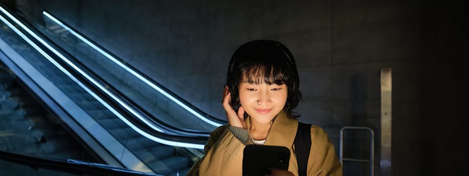 Portrait of beautiful korean girl in headphones, stands near escalator, travels, commutes home on public transport, holds smartphone, poses with mobile phone.