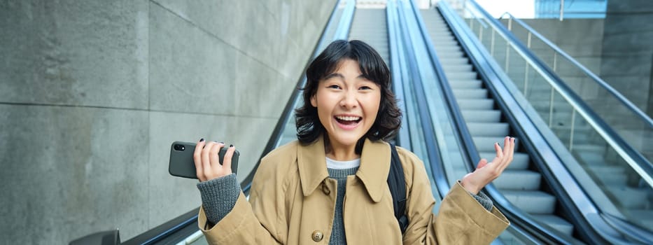 Happy triumphing asian girl, looks amazed and joyful, holds smartphone, goes down escalator with pleased face, wins, hears good news.