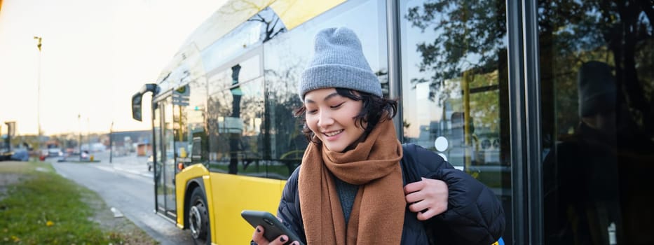 Image of girl student waiting for public transport, checks schedule on smartphone app, stands near city bus. Urban lifestyle concept